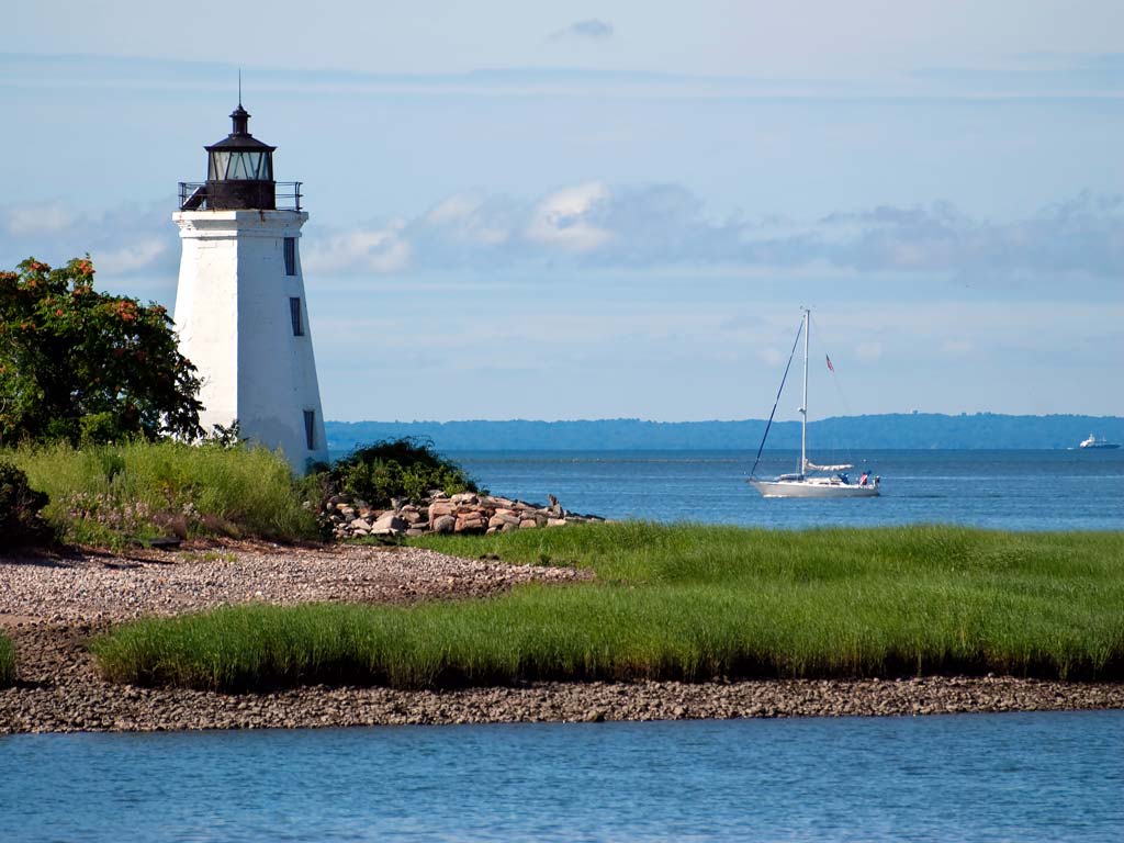 A photo of the Fayerweather Island Light, a lighthouse located on the shores of Bridgeport, Connecticut, with a sailboat passing by the coast
