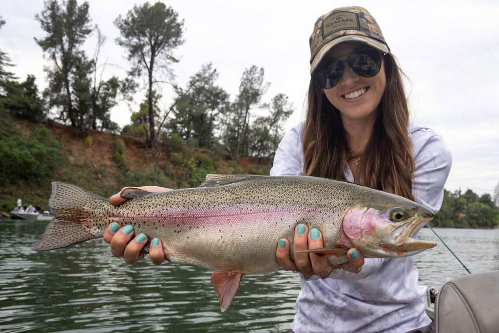 A woman in a hat and sunglasses smiling as she holds a big Rainbow Trout towards the camera, the fish is big but nowhere near the size of the California state record Trout.