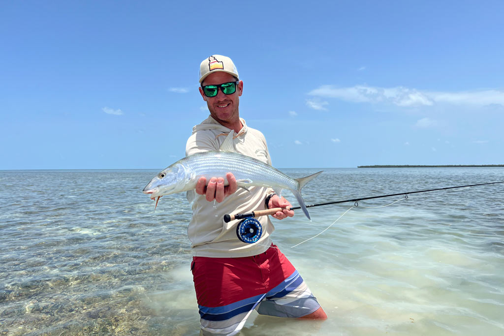 A photo featuring an angler standing in the shallows and posing with a fly fishing rod in one hand and a Bonefish in the other hand