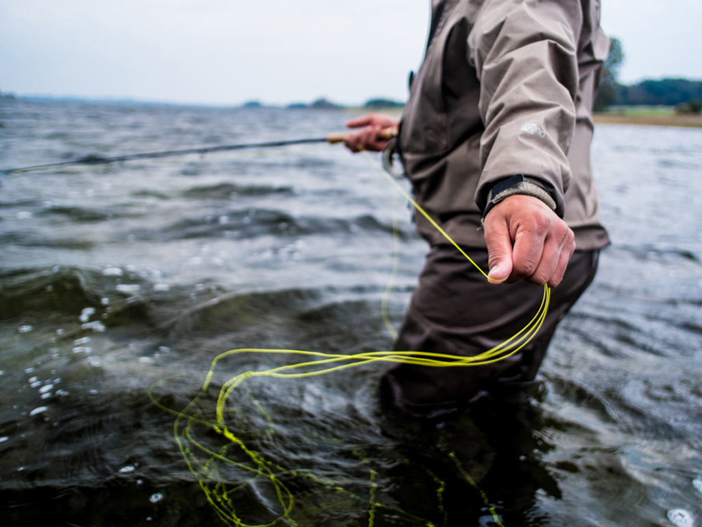 An image of a fly fisherman from the neck down standing waist-deep in water and pulling line towards him