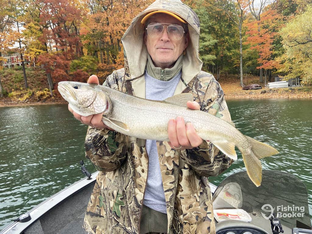 A photo featuring an older angler standing on a charter boat while calmly posing with a Trout he caught on a lake surrounded by scenic fall foliage