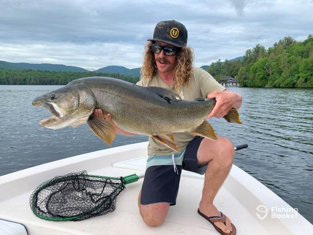 A photo featuring an angler on a charter boat posing with a decent Lake Trout catch on a cloudy but warm spring day