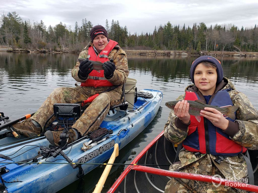 A photo featuring a father and a son on a cold day during the fall fishing season while they’re sitting in a blue and red kayaks and posing with a catch each
