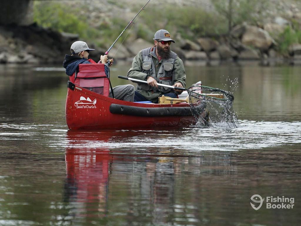 A photo featuring a father and a son sitting in a kayak and angling for Trout on a river with a rod and net on a cold but great day for angling
