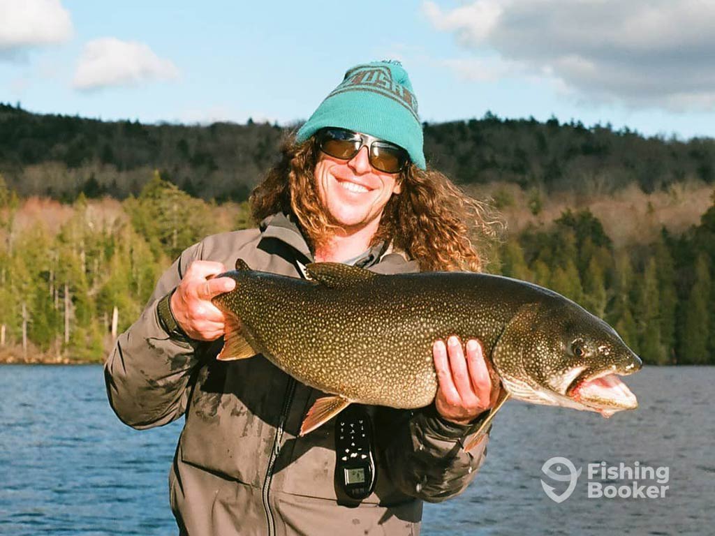 A photo featuring a proud angler standing on a charter boat in the middle of a lake and posing with a decent Lake Trout caught during the fall fishing season