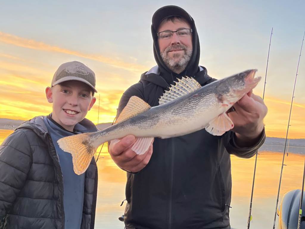 A man and a boy standing on a fishing charter on a river or a lake, as the man holds a Sauger with the setting sun visible behind them across the water