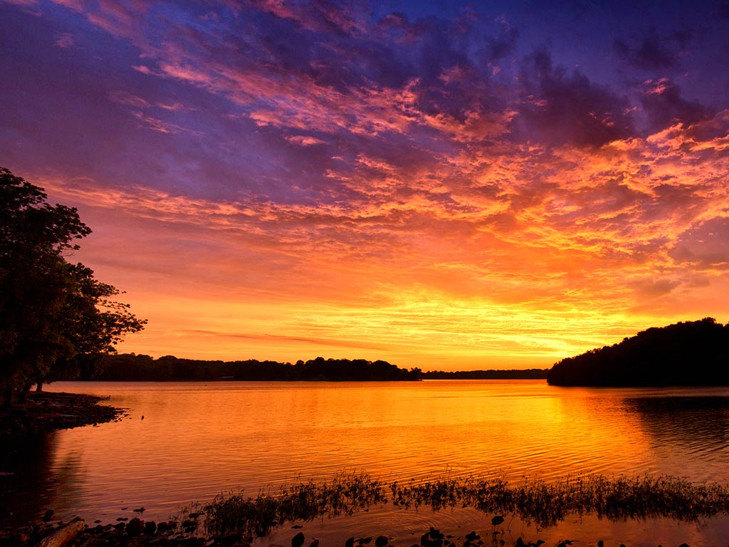 A view across Old Hickory Lake in Tennessee at sunset, with an island in the river visible on the right of the image and the sun setting in the distance, creating orange and red hues in the sky