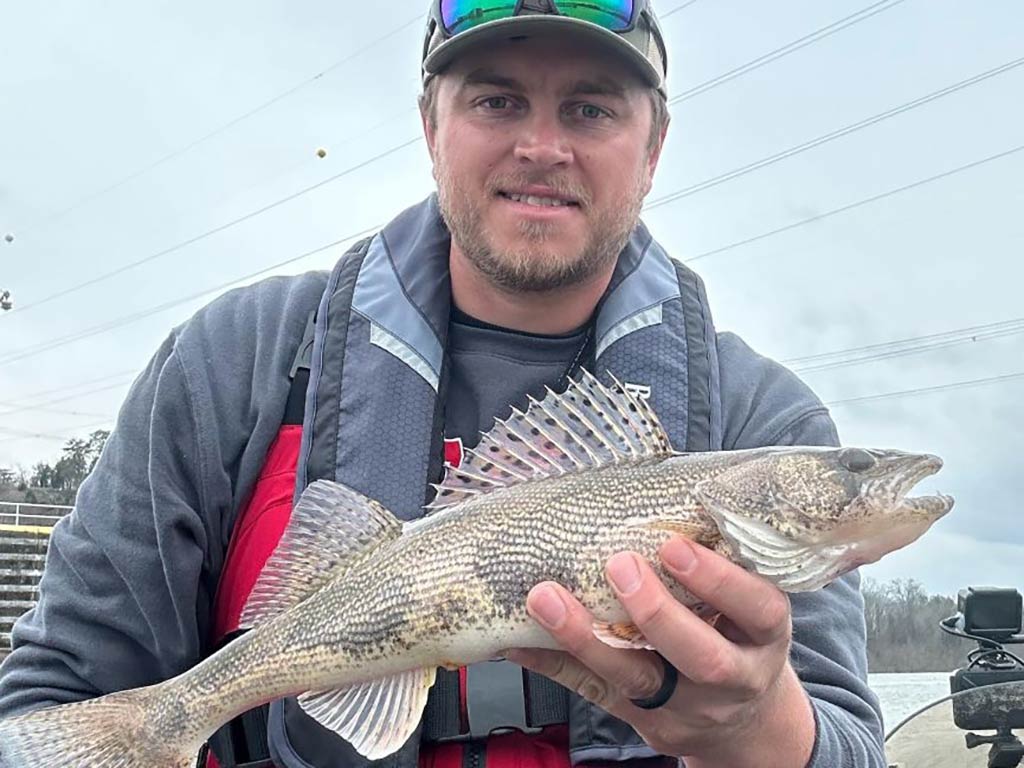 A man with sunglasses on his baseball cap wearing a life vest and holding a colorful Sauger to the camera on an overcast day