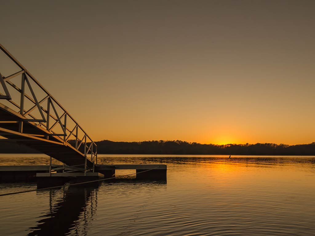 A view across Dale Hollow Lake at sunset, looking towards the sun setting on the opposite bank of the lake, with a boat launch visible on the left of the image