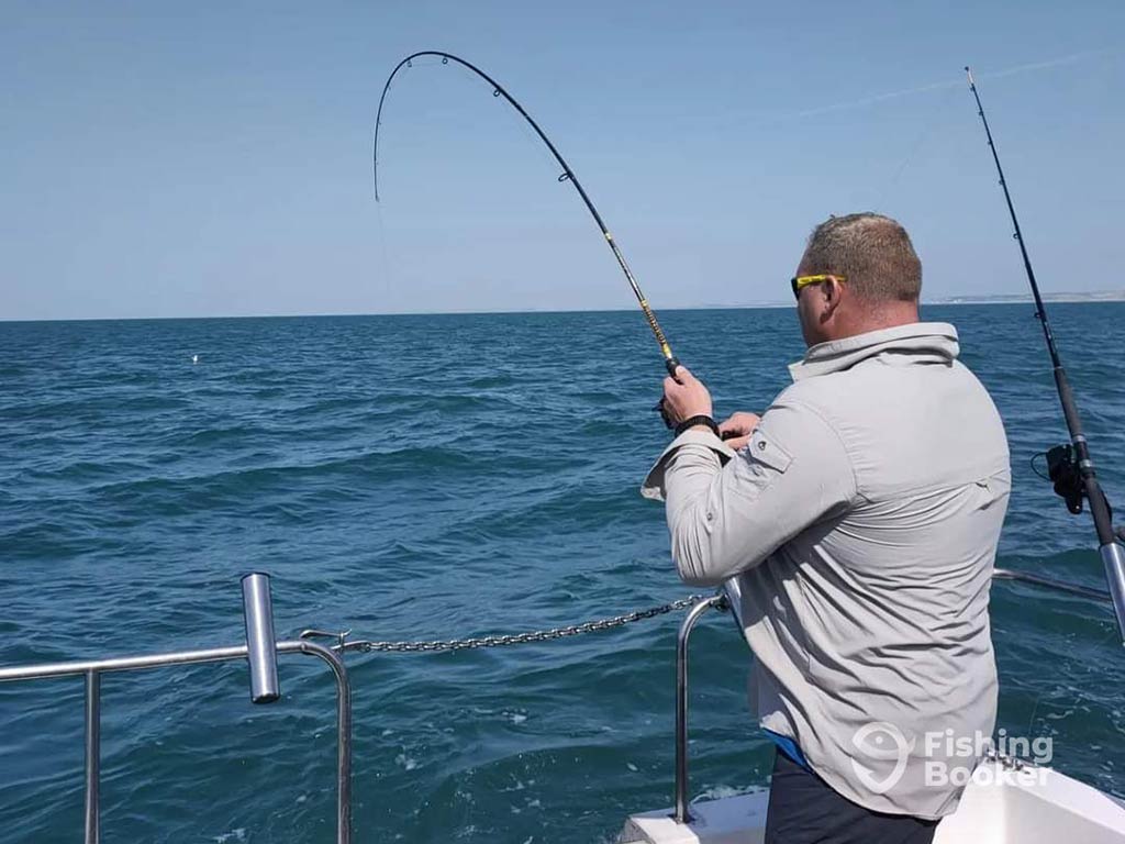 A view from behind of a man fishing over the side of a boat in the UK on a clear day, with another bent rod visible next to him