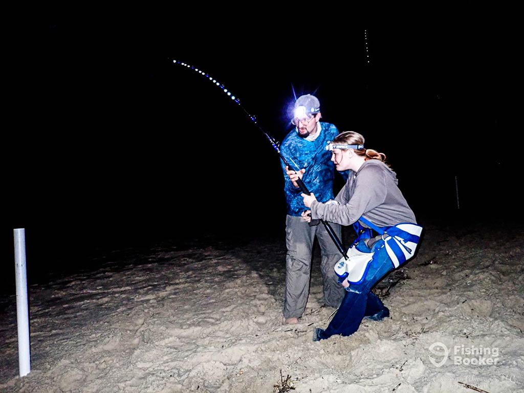 A woman crouching and struggling while fishing at night on a beach as a male angler helps her