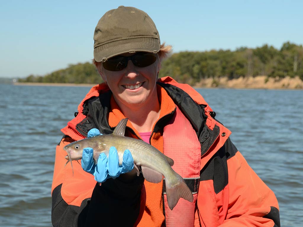 A man in an orange raincoat, hat, and sunglasses holding a White Bullhead next to a river or lake on a  clear day
