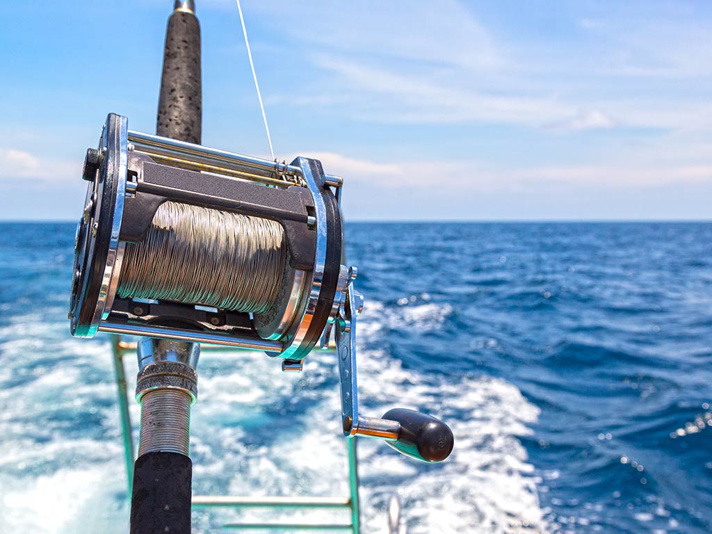 A closeup of a heavy-duty fishing reel spooled with a wire line ready for deep sea fishing, with open ocean waters and the wake of a boat visible behind the reel on a sunny day