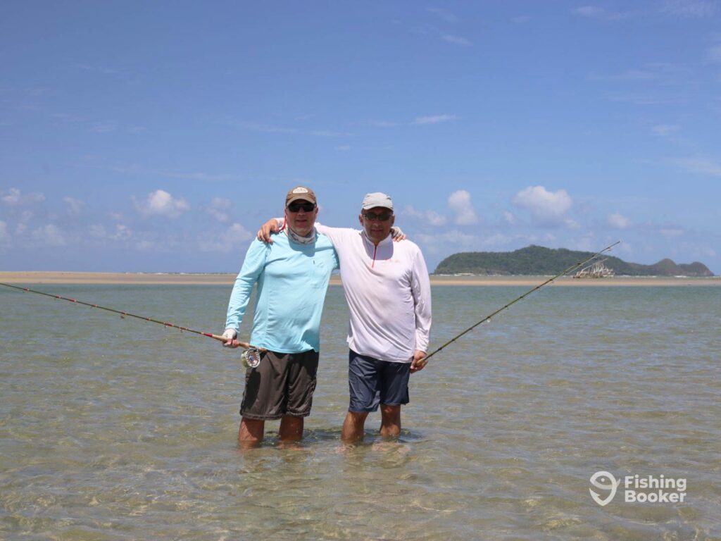 Two middle-aged men wading in the crystal clear shallow flats in Cairns and holding a fly fishing rod each on a clear day