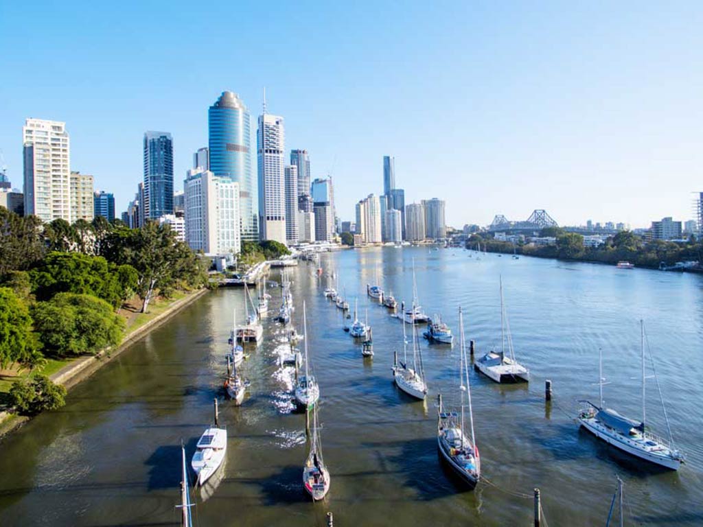 An aerial view looking down a river lined with boats docked towards the skyline of Brisbane on a clear day