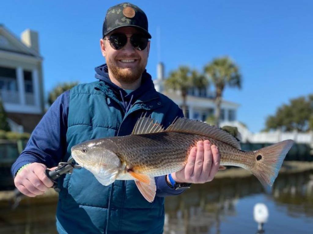 A photo featuring a smiling angler wearing a pair of sunglasses and a cap while posing on a charter fishing boat in front of houses with a decent Redfish caught in Myrtle Beach around Thanksgiving