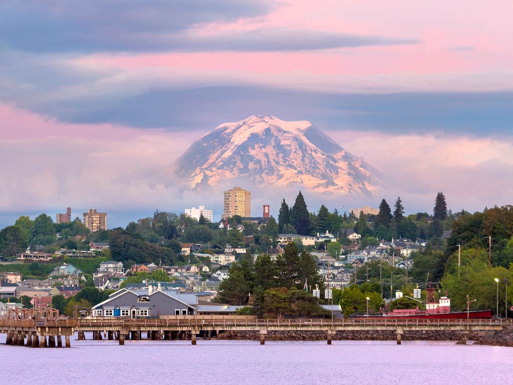 A magnificent view of the town of Tacoma and Mount Rainier behind it seen from the water during the sunset hour full of violet and pink skies and hues