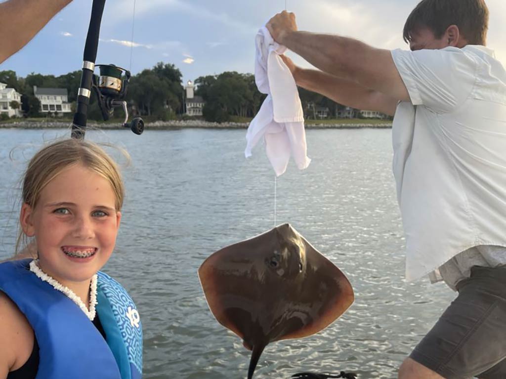 A little girl smiles as she looks towards the camera, while a man carefully handles a Ray on the end of a fishing line behind her on a boat