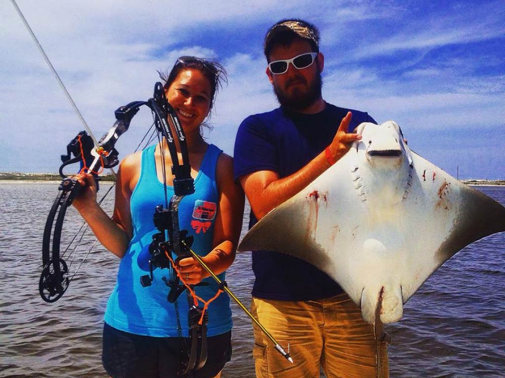 A man and woman standing on a boat, as the man holds a Stingray and the woman a bowfishing gun on a clear day