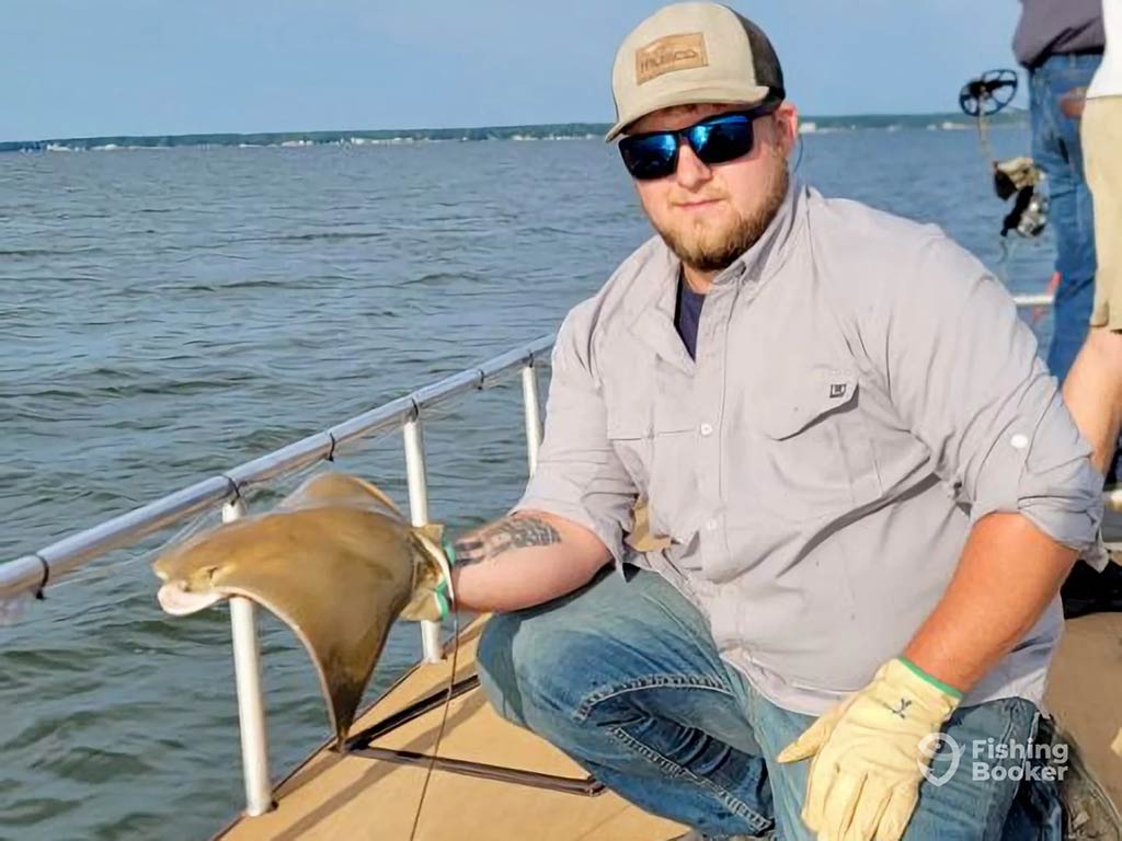 A man in sunglasses and a baseball cap poses with a Ray flapped over his hand, while wearing gloves aboard a fishing boat