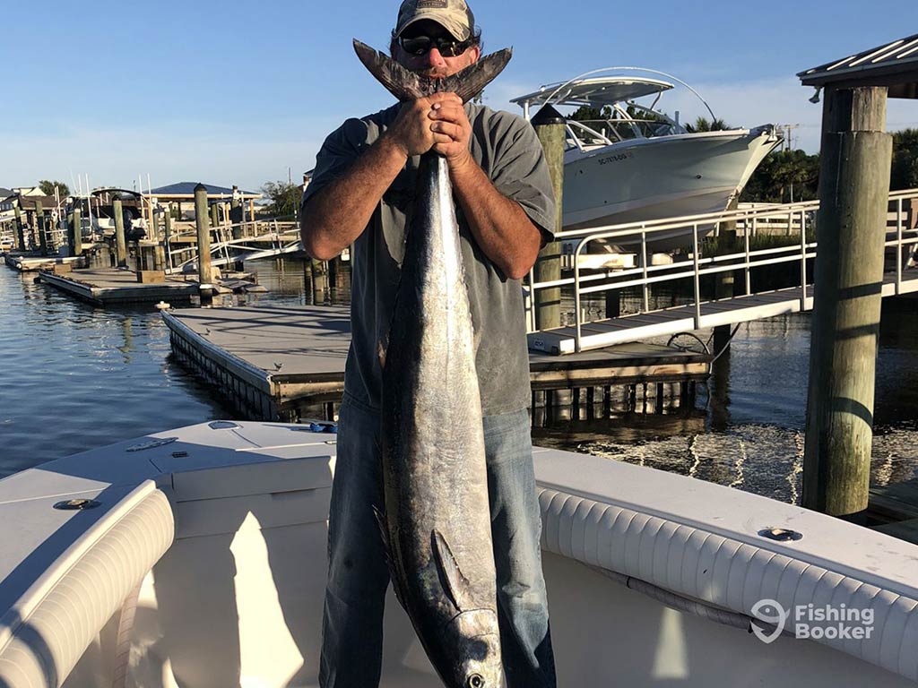 A man in a hat and sunglasses standing on a dock in Hilton Head Island and holding a large Wahoo vertically up above his face