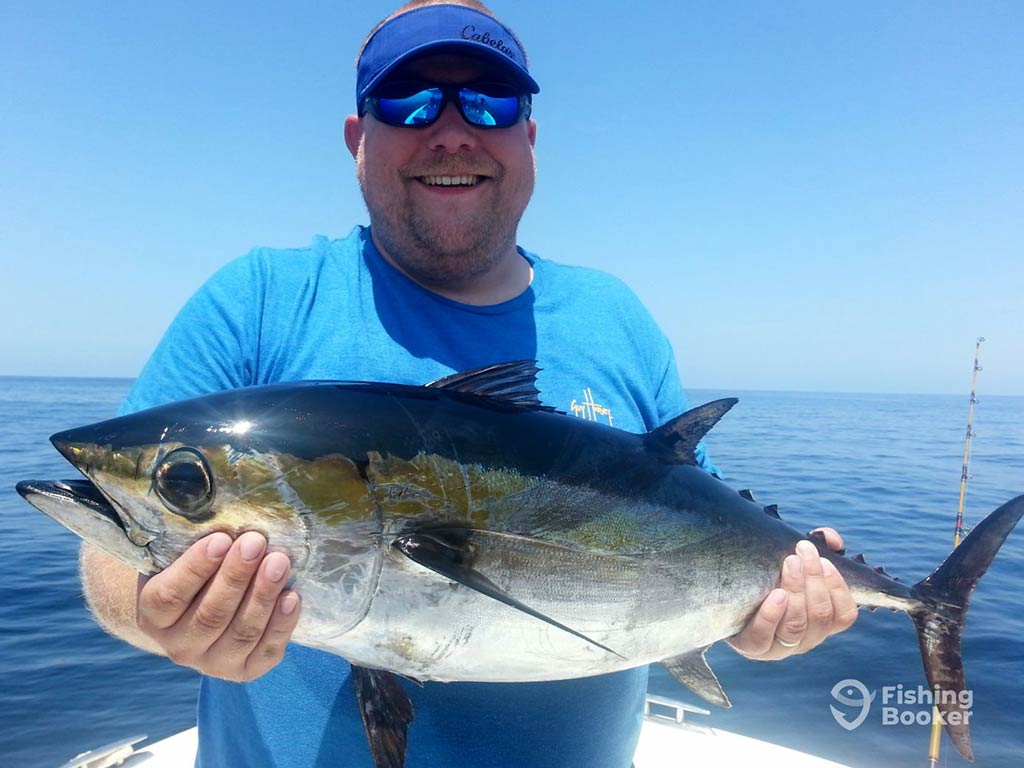 A man in a baseball cap and sunglasses smiling as he holds a Blackfin Tuna aboard a fishing charter in the Atlantic Ocean on a clear day