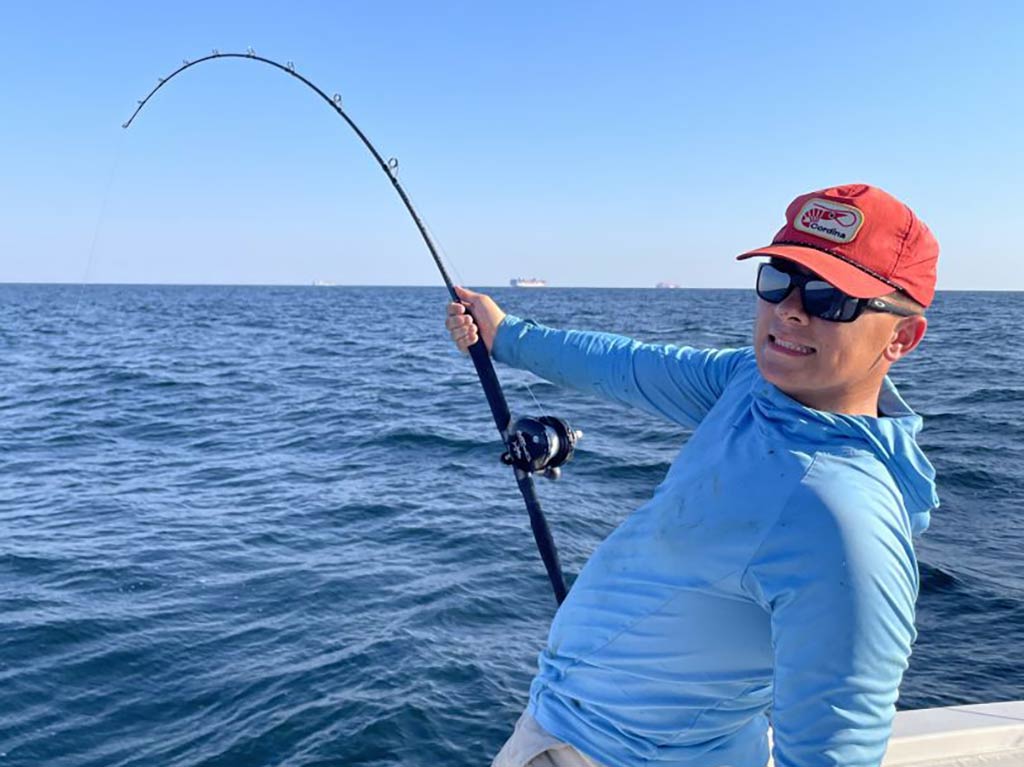 A man in a red baseball cap and sunglasses bottom fishing over the side of a fishing charter out of Hilton Head as he looks back towards the camera on a clear day
