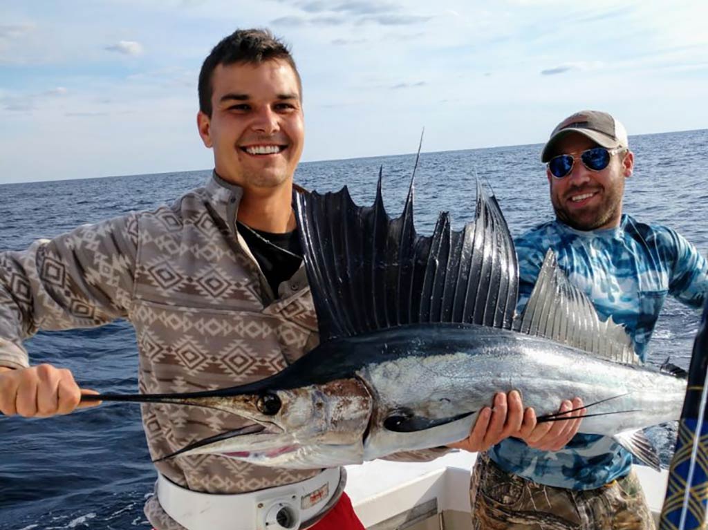 Two men standing on a fishing charter out of South Carolina and holding a Sailfish between them on a bright day