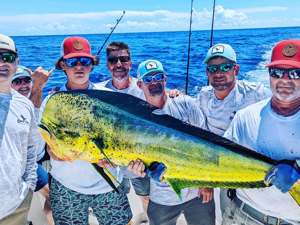 A group of male anglers aboard a fishing charter smiling as they hold a large Mahi Mahi between them on a clear day