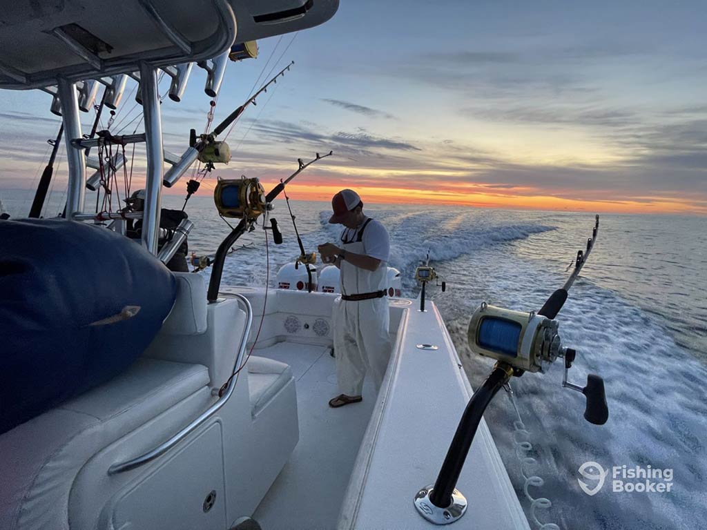 A view along the side of a center console boat towards the deck, where a captain or first mate is unrigging a fishing rod with other trolling rods visible at sunset on the open waters