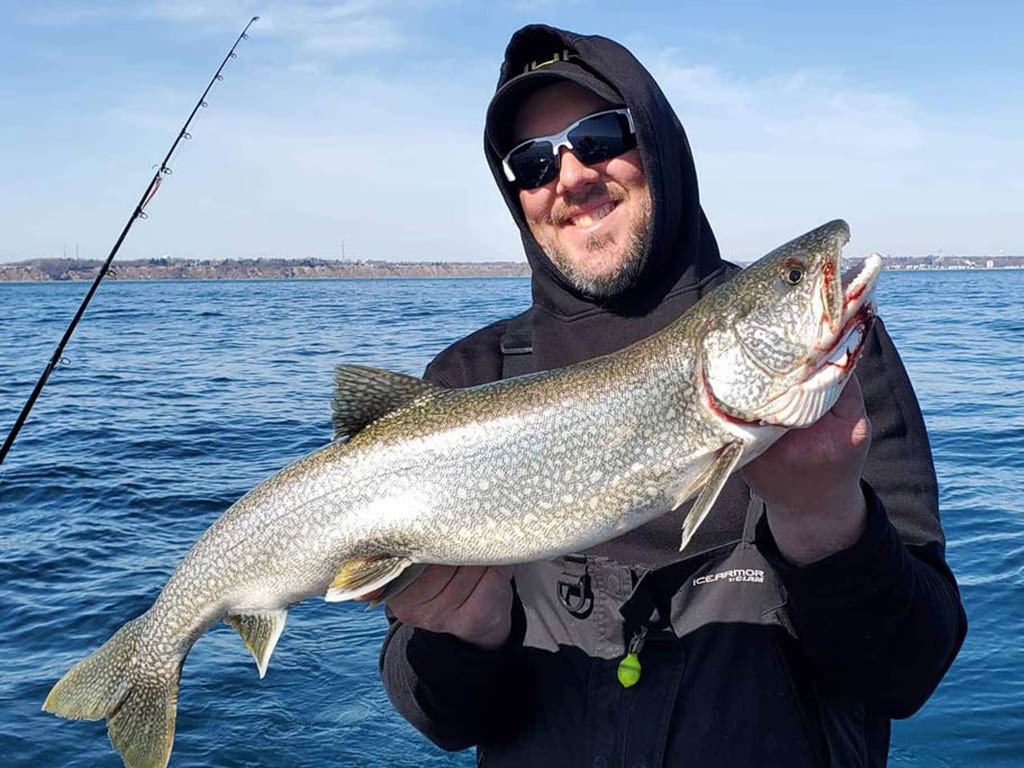 A smiling fisherman in a hoodie and sunglasses, standing on a fishing boat, holding a big Lake Trout, with blue skies and water in the background