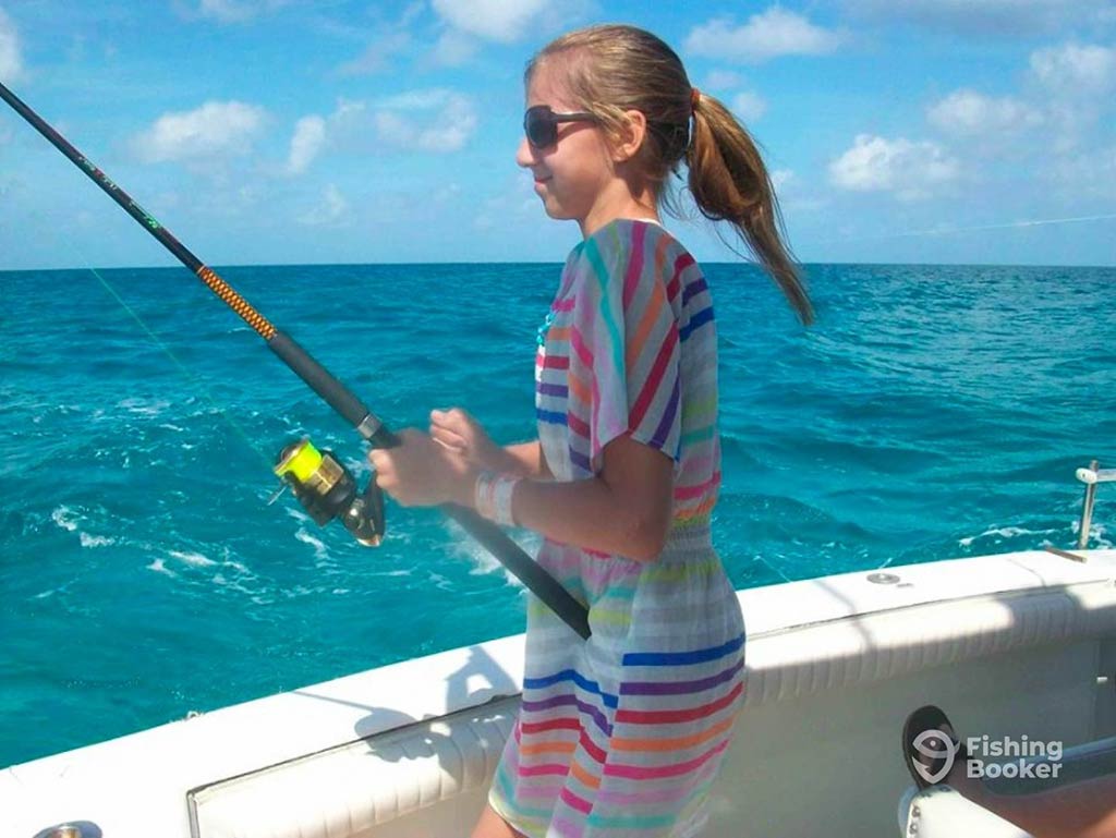 A young woman holding a fishing rod with both hands aboard a fishing charter in the Bahamas, with clear waters visible behind her on a bright day