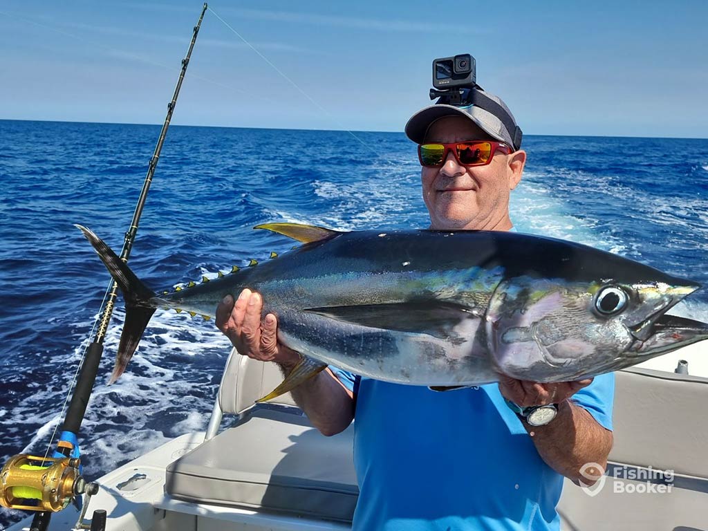 A man wearing a baseball cap with a GoPro on top of it smiling as he holds a Blackfin Tuna aboard a deep sea fishing charter in the Bahamas on a sunny day