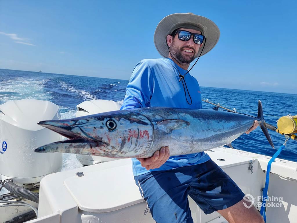 A man in a Panama hat smiling as he sits on the side of a fishing charter on a clear day, holding a large Wahoo
