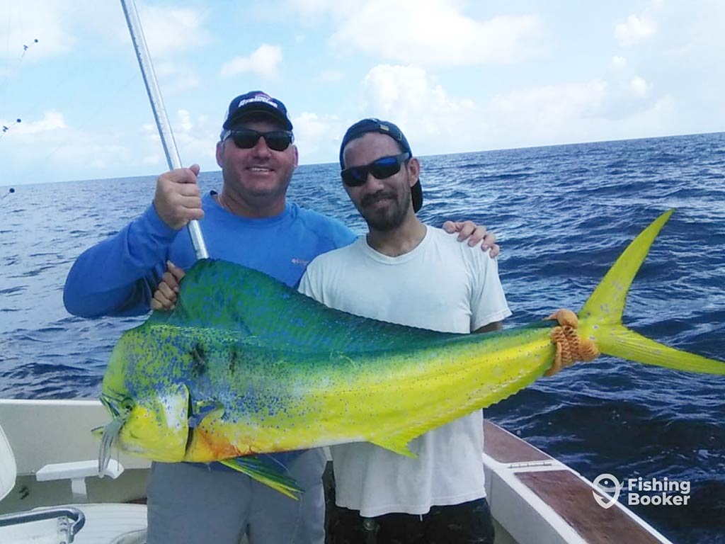 Two men in sunglasses standing aboard a fishing charter and showing off their large Mahi Mahi catch with deep waters visible behind them
