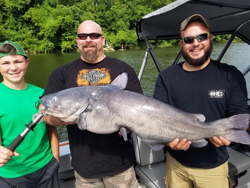 A photo featuring three anglers, two adults and a kid, standing on a charter boat and smiling while posing with a Blue Catfish they caught after learning how to catch Catfish