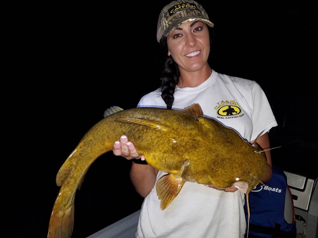 A smiling woman wearing a cap and white T-shirt while standing on a charter boat at night and holding a Flathead Catfish in both hands, with nothing but darkness behind her.