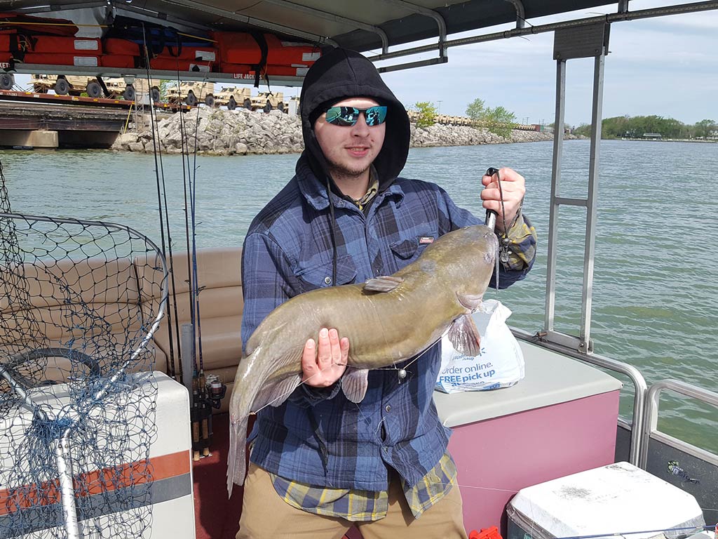 A photo of an angler standing on a charter boat in sunglasses and his hood on while holding a Channel Catfish with a fishing net on the left and water on the right