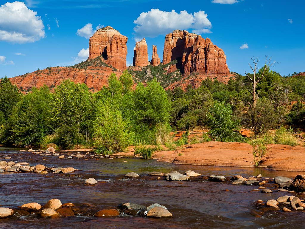 A view looking up towards rock formations on the top of a rocky mountain in Sedona, AZ, on a sunny day with some greenery in the foreground