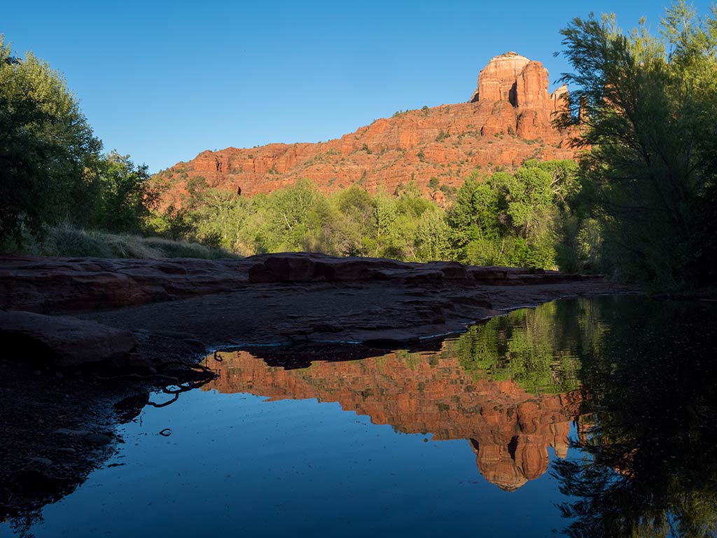 A view towards Cathedral Rock in Sedona, AZ, from Oak Creek with calm waters in the shadow in the foreground and blue skies in the distance