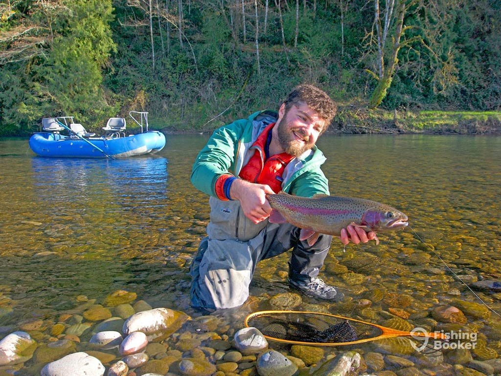 A man in waterproof gear crowching down in a clear river and posing with a Rainbow Trout in his hands with an inflatable blue boat visible across the water behind him