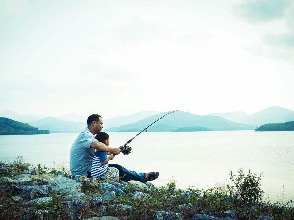 A father and son sitting on a hill overlooking a lake on a cloudy day, as they fish with the same rod