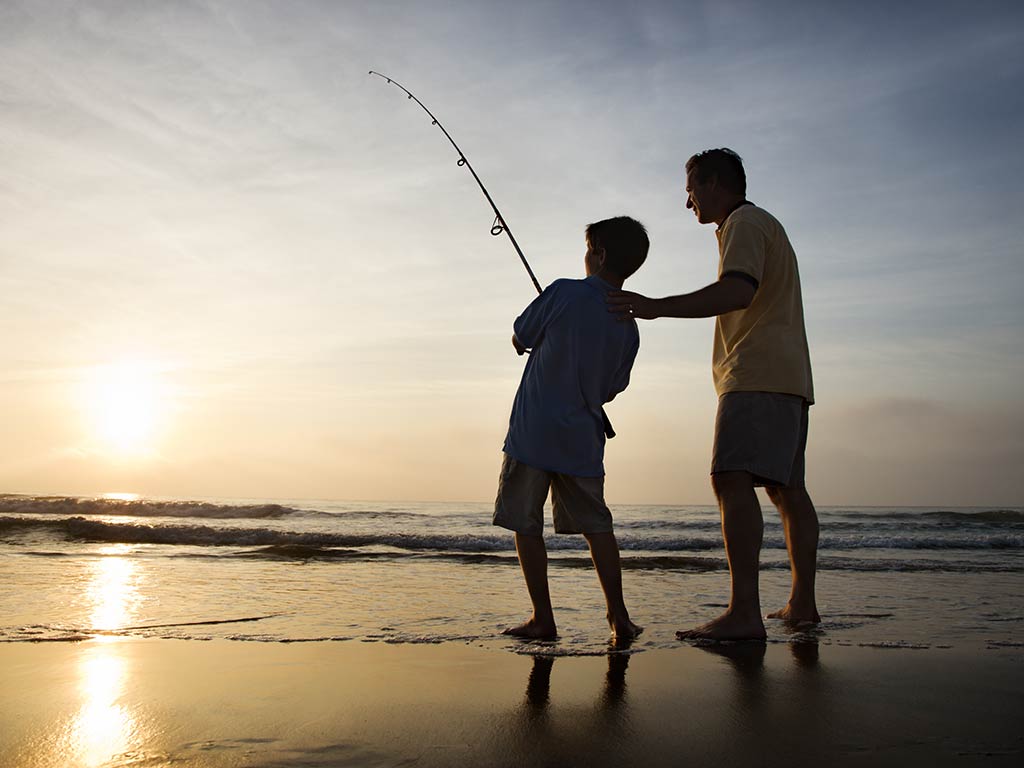 A view from behind of a father and son fishing on a beach at sunset on a clear day