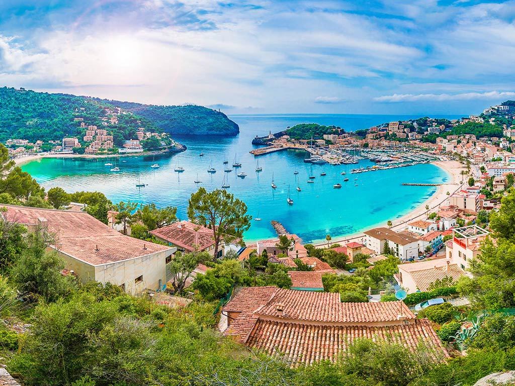 A view from a hill looking towards a harbor on the island of Mallorca, with crystal clear waters visible in the bay and an inlet leading to the open waters of the Mediterranean beyond it