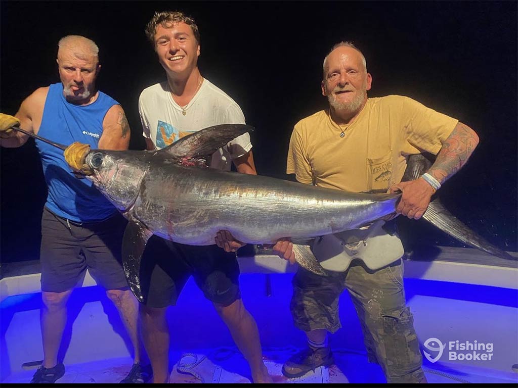 Three men holding a large Swordfish aboard a deep sea fishing charter at night