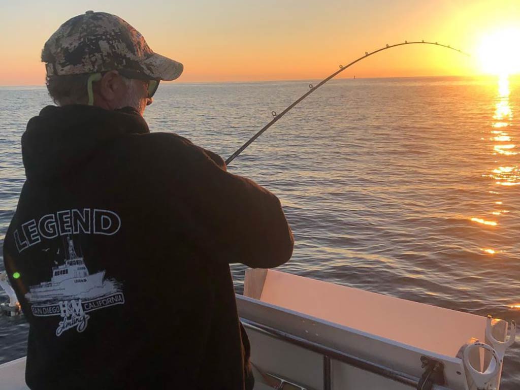 A view from behind of an angler in a hoodie and baseball cap jigging in the waters off California's coast at sunset, with the sun visible on the right of the image