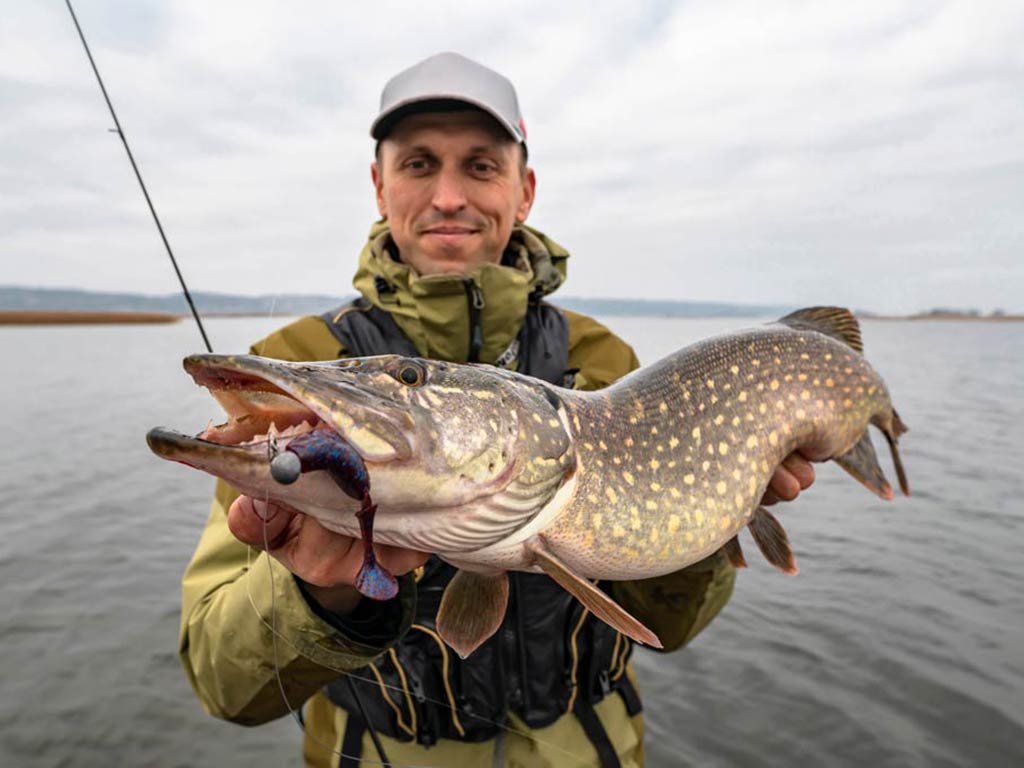 A man in a baseball cap, and a khaki winter jacket posing next to a lake while holding his fishing rod and a large Northern Pike with a jig in its mouth on a cloudy day
