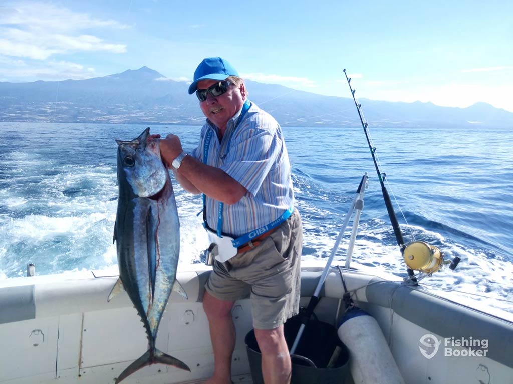 A man in a blue baseball hat and sunglasses standing on a fishing charter in the Mediterranean and holding a Bigeye Tuna on a clear day with a mountain visible in the distance across the waters