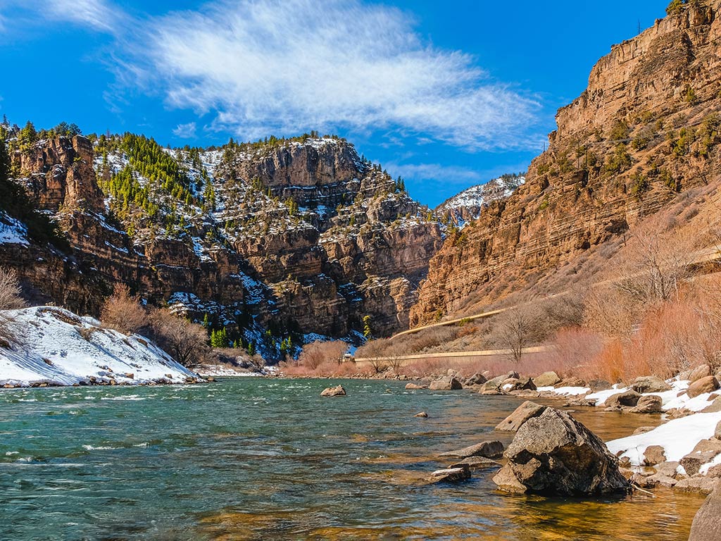 A view looking down the Colorado Rover near Glenwood Springs on a clear day, with large rocky mountains all around the water.