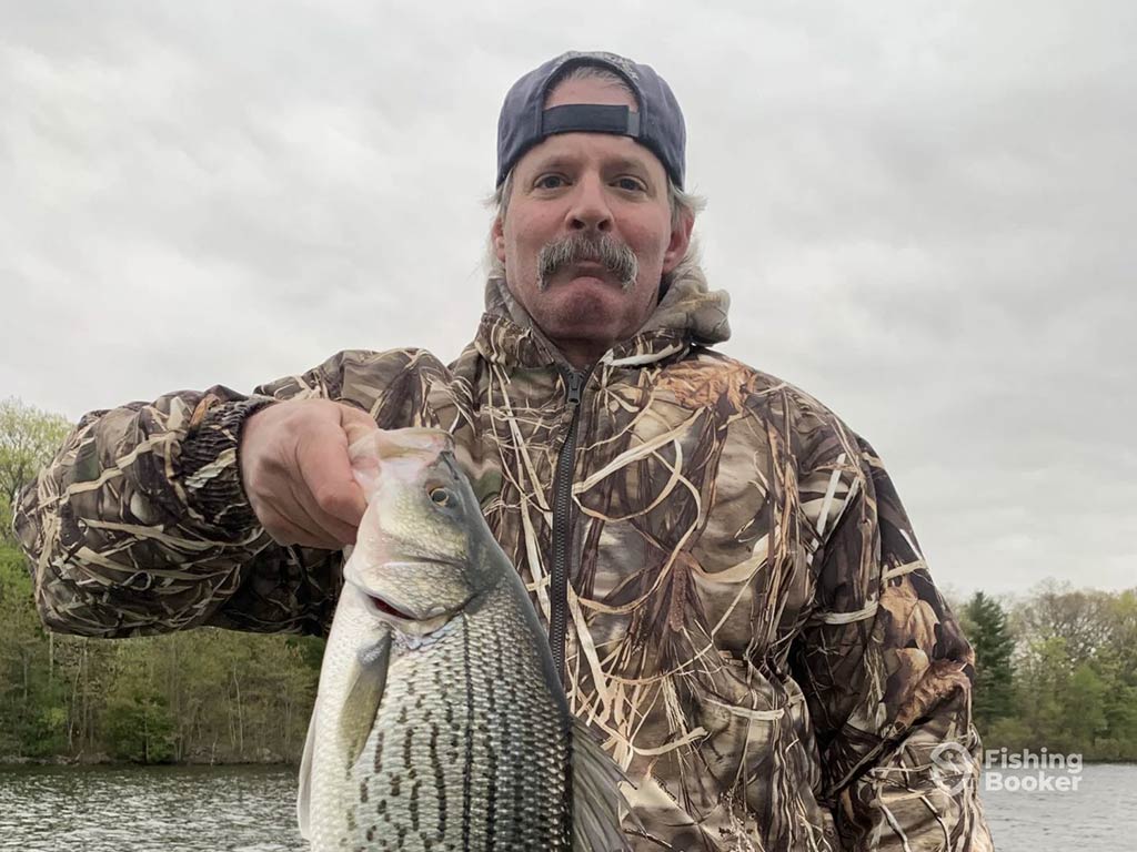 A man in a back-to-front baseball cap and camouflage coat holding a Hybrid Striped Bass on a lake near Hopatcong, NJ, on a cloudy day